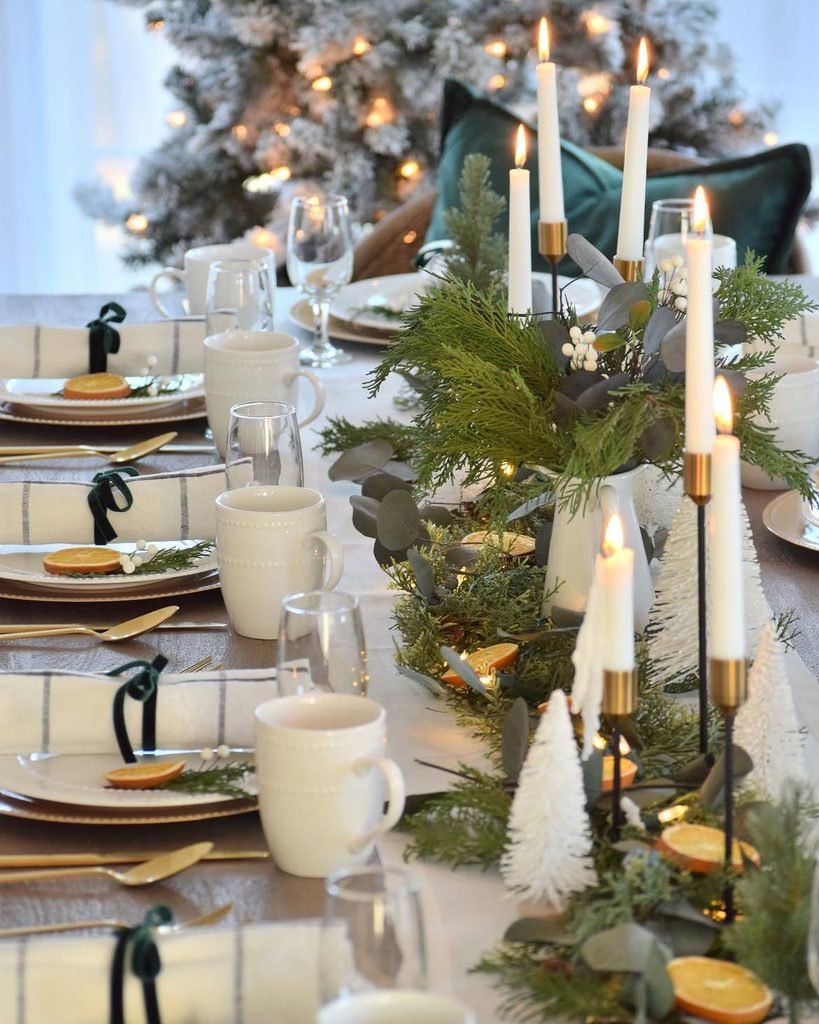 Dining table decorated with three small Christmas trees, cream tableware, and natural greenery