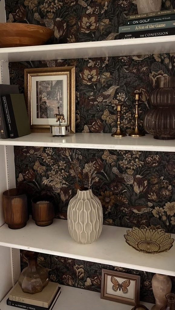 Dark green wallpaper behind built-in shelves with white ceramics, books, and brass accents